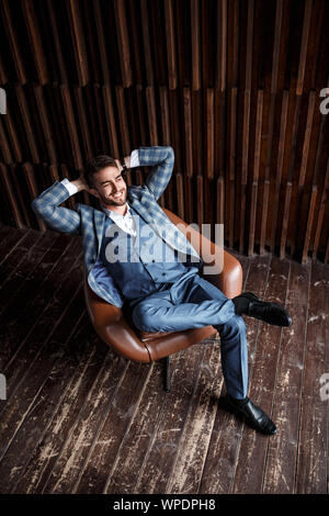 Junge erfolgreiche Geschäftsmann in einem blauen Anzug in einem Käfig sitzt in einem Ledersessel. junger Mann mit einem Bart lächelnd Stockfoto