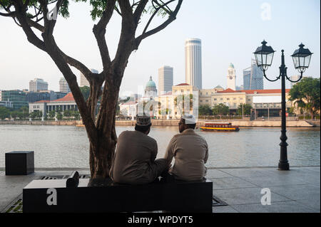 07.09.2019, Singapur, Republik Singapur, Asien - Zwei muslimische Männer sitzen auf einer Bank am Boat Quay Entlang des Singapore River an der Skyline der Stadt. Stockfoto