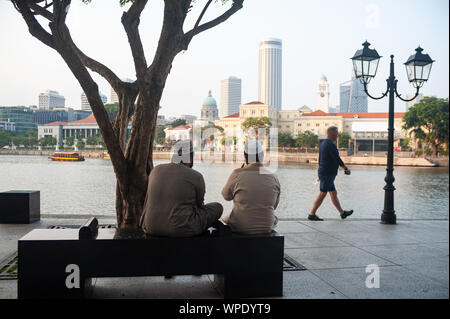 07.09.2019, Singapur, Republik Singapur, Asien - Zwei muslimische Männer sitzen auf einer Bank am Boat Quay Entlang des Singapore River an der Skyline der Stadt. Stockfoto