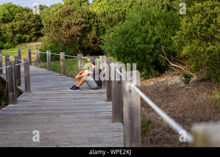 Bärtiger Mann trägt eine Kappe in einem Abstand sitzen auf dem Boden eines Holz- weg von der Natur umgeben. Stockfoto