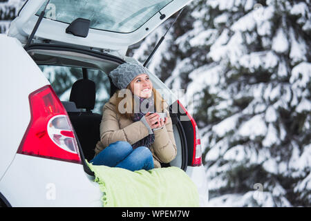 Junge Frau traveler sitzt in den Kofferraum des Autos und hält eine Tasse heißen Tee in den Händen. Stockfoto