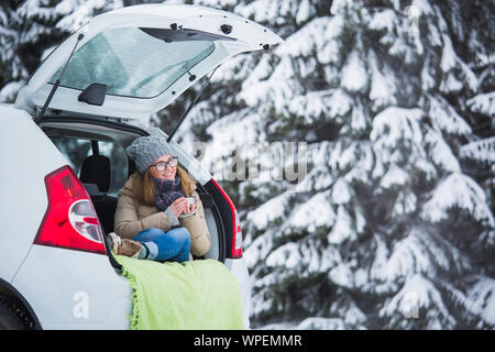 Junge Frau traveler sitzt in den Kofferraum des Autos und hält eine Tasse heißen Tee in den Händen. Stockfoto