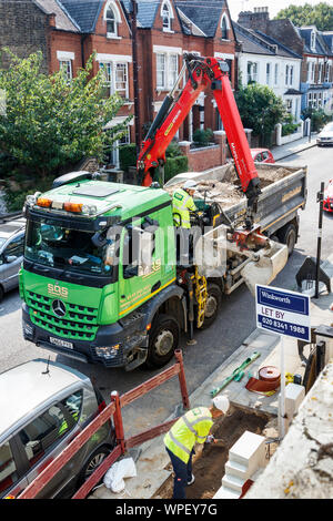 Zwei männliche Arbeitnehmer in hohe Sichtbarkeit Jacken und Mützen, einer Steuerung der Grabber auf einem Lkw, die anderen füllen ein Loch im Bürgersteig, London, UK Stockfoto