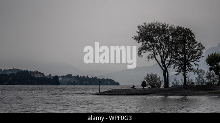 Ein graues nebligen Morgen am See Maggiori in Italien. Eine Person sitzt auf einer Bank an einem kleinen See der Insel, die über den Sturm kalten Blick. Stockfoto