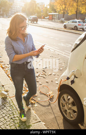 Junge Frau steht in der Nähe des elektrischen Auto und schaut auf dem Smartphone. Der mietwagen wird der Ladevorgang an der Ladestation für Elektrofahrzeuge. Stockfoto