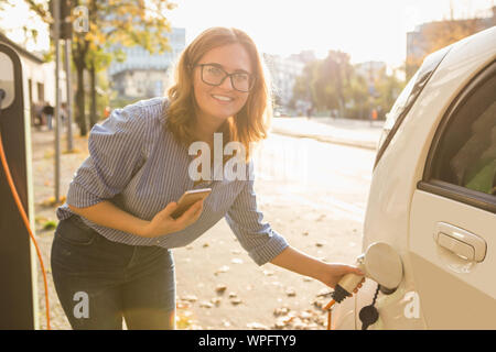 Junge Frau steht in der Nähe des elektrischen Auto und schaut auf dem Smartphone. Der mietwagen wird der Ladevorgang an der Ladestation für Elektrofahrzeuge. Stockfoto