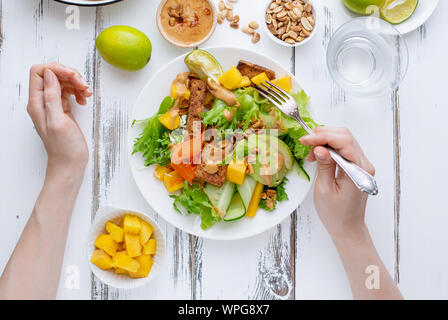 Weibliche Hände halten einen Teller mit frischem Salat mit Mango, Avocado, Kalk und Tofu mit süßen Peanut butter Dressing weiße Holztisch Stockfoto