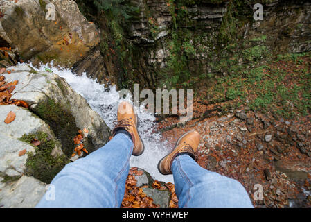 Mann Beine in Jeans und braune Stiefel sitzen auf der Flanke am Wasserfall suchen Stockfoto