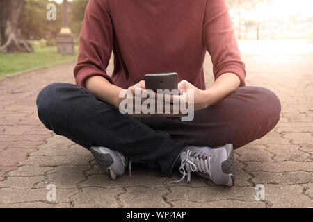 Hände halten ein Smartphone und auf dem Boden sitzend den Sonnenuntergang im Garten. Stockfoto