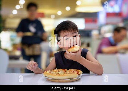 Asiatische 6-7 Jahr Junge ist glücklich essen Pizza mit einem heißen Käse auf einem hölzernen Pad in Restaurant gestreckt zu schmelzen. Stockfoto