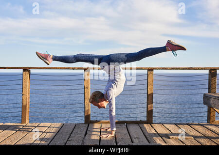 Volle Länge senior Frau macht Handstand während des Trainings auf Diele auf See gegen Sky Stockfoto