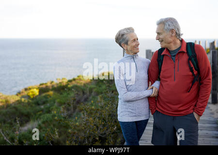 Lächelnd senior Paar mit Arm in Arm mit Blick auf die jeweils andere beim Stehen auf der Promenade von Sea gegen Sky Stockfoto