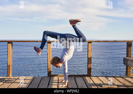 Volle Länge senior Frau macht Handstand während des Trainings auf Diele auf See gegen Sky Stockfoto