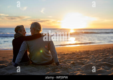 Ansicht der Rückseite des älteren Ehepaar miteinander reden während der Sitzung auf Sand am Strand bei Sonnenuntergang Stockfoto