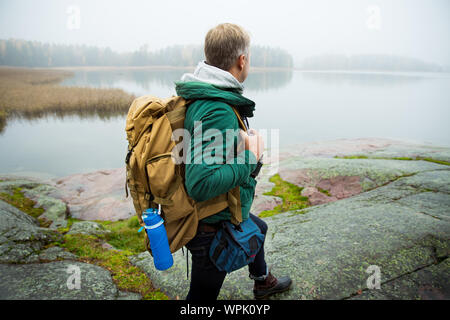 Reifer mann Finnland erkunden im Herbst, in Nebel. Wanderer mit grossen Rucksack auf bemoosten Felsen. Skandinavische Landschaft mit misty Meer und Stockfoto