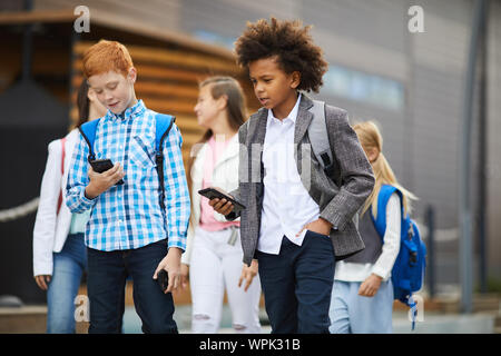 Zwei Schuljungen mit Rucksäcken zu Fuß entlang der Straße und sprechen über Mobiltelefone mit ihren Eltern aus der Schule kommen Sie zurück Stockfoto