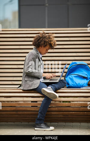 Afrikanische Schüler auf der Bank sitzen allein und Sie auf seinem Laptop Computer er seine Hausaufgaben im Freien tun Stockfoto
