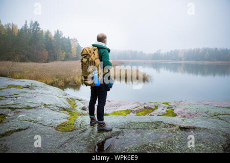 Reifer mann Finnland erkunden im Herbst, in Nebel. Wanderer mit grossen Rucksack auf bemoosten Felsen. Skandinavische Landschaft mit misty Meer und Stockfoto