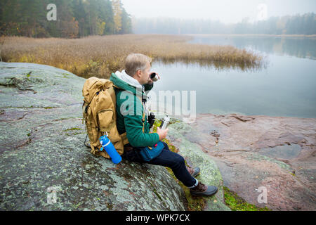 Reifer mann Finnland erkunden im Herbst. Wanderer mit grossen Rucksack sitzen auf bemoosten Felsen, trinken heißen Kaffee aus der Thermoskanne. Skandinavische Landschaft mit Stockfoto