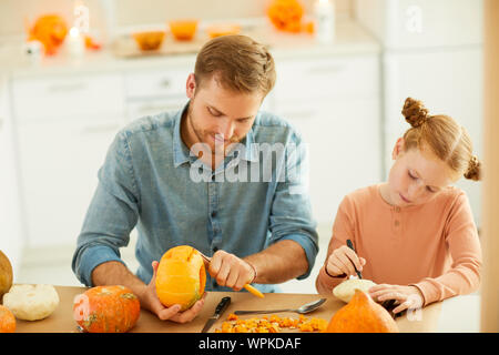 Vater Peeling der Haut eines Kürbis mit kleinen Mädchen Zeichnung etwas auf Sie, während sie am Tisch in der Küche zu Hause sitzen Stockfoto