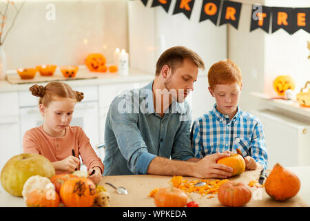Junger Vater am Tisch sitzen zusammen mit seiner Tochter und Sohn und Ihnen hilft, Jack O'Lantern von Kürbissen zu machen Stockfoto