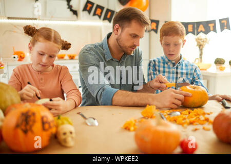 Junger Vater am Tisch sitzen zusammen mit seinen zwei Kindern und sie vorbereiten Kürbisse für Halloween Party zu Hause Stockfoto