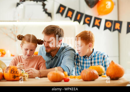 Süße Mädchen und junge Dekorieren reife Kürbisse, die zusammen mit ihrem Vater am Tisch zu Hause sitzen Stockfoto