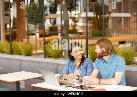 Junge lächelnde Frau mit bärtiger Mann, während Sie am Tisch vor Laptop im Café draußen sitzen Stockfoto