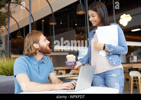 Glückliche junge Frau, Kaffeegetränke, junger Mann, während er am Tisch sitzen und im Laptop Computer sie Ausgabe Zeit eingeben Stockfoto