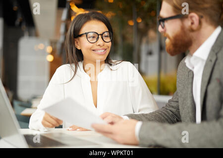 Junge erfolgreiche Geschäftsfrau in Brillen am Tisch sitzen zusammen mit Geschäftsmann und sie diskutieren Vertrag zusammen Stockfoto