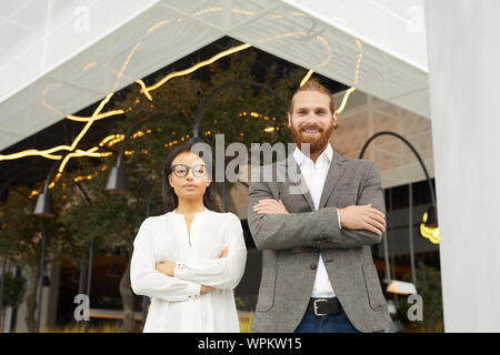 Porträt der Bärtige junge Unternehmer Stehen mit verschränkten Armen und lächelnd an Kamera mit selbstbewussten Geschäftsfrau steht in der Nähe von ihm im Freien Stockfoto