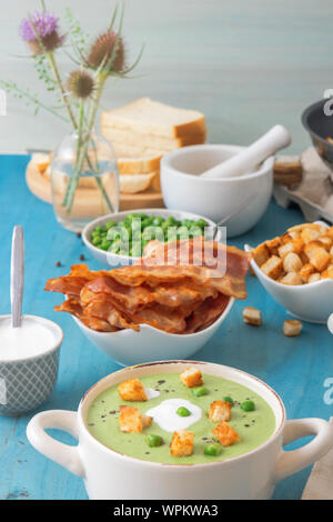 Blau Holztisch mit Schalen der Dicke grüne Erbsensuppe mit gerösteten Zwieback, grüne Erbsen verschönert, Scheiben mit gegrilltem Speck, Zerkleinerter schwarzen Pfeffer und cre Stockfoto