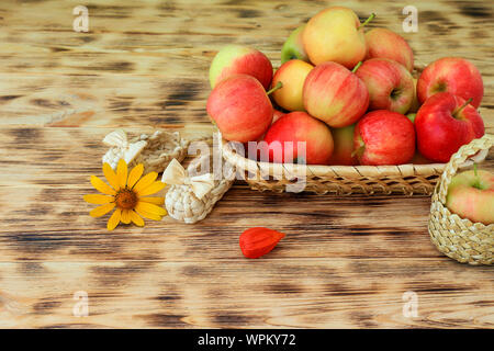 Rote Äpfel und Herbst Blumen liegen in einem wicker Platte auf einem Holztisch. Wicker bast Schuhe und Äpfel in einem Korb. Gesunde Ernährung und Lebensstil Stockfoto