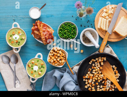 Blau Holztisch mit Schalen der Dicke grüne Erbsensuppe mit gerösteten Zwieback, grüne Erbsen verschönert, Scheiben mit gegrilltem Speck, Zerkleinerter schwarzen Pfeffer und cre Stockfoto