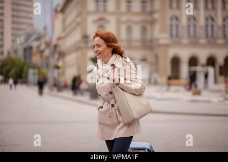 Ein Lächeln auf den Lippen reife Frau, Wandern in einer Fußgängerzone, in einer Stadt. Reisende ihren Koffer zu schieben. An einem bewölkten Tag im Freien. Stockfoto