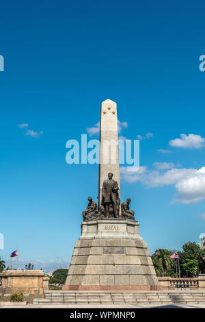 Manila, Philippinen - 5. März 2019: Obelisk mit Bronze Statuen von Jose Rizal steht auf braunem Stein Podest unter blauem Himmel in Rizal Park. Fahnen und Stockfoto