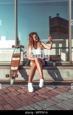 Schöne Mädchen mit Brille, in der Nähe von Gebäude sitzen, im Sommer in der Stadt, der Tasse Kaffee, Fotos selbst am Telefon, lächelt glücklich, Video Stockfoto