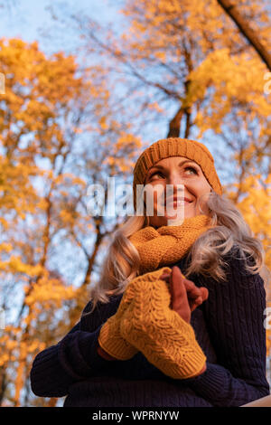 Ein Mädchen steht im Herbst im Oktober auf einer weißen Plattform in einem Wald, in einem Hut, in Fäustlingen, in einer schwarzen Jacke gegen einen strahlend blauen Himmel Stockfoto