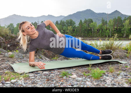 Blonde schöne Mädchen lächelnd und ein Warm-up liegen auf dem Stein- Boden in der Pose des seitlicher Ganzkörperstütz, stützte sich auf den Ellenbogen draußen in den mounta Stockfoto