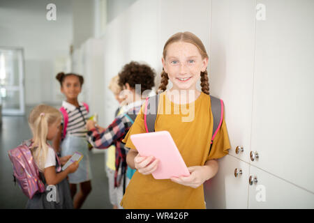 Mädchen mit Rucksack Holding rosa Tablette beim Stehen in der Nähe von Schließfächern Stockfoto