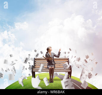 Frau mit offenem Buch auf Holzbank draußen. Mädchen in Business Suit Finger, der auf Sky. Panorama mit runden Horizont der Welt und fliegen Papier doc Stockfoto