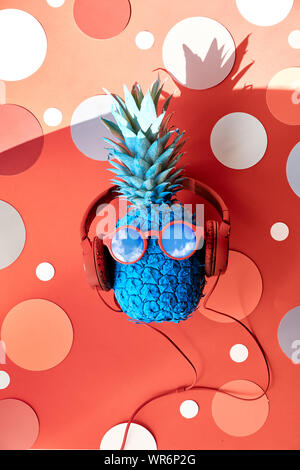Lustige Ananas blau lackiert in Rot in Sonnenbrille und Kopfhörer. Flach auf Coral Farbe Papier mit Kreisen. Stockfoto
