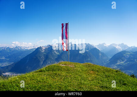 Österreichische Fahne im Wind zu einem Gras bedeckte Gipfel mit klaren, blauen Himmel und die Alpen im Hintergrund an einem Sommertag Stockfoto