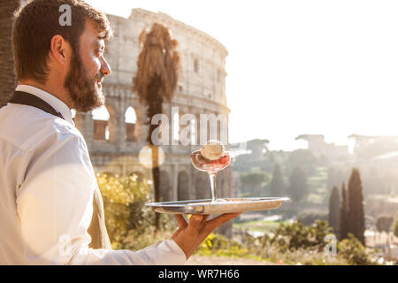 Elegante kellner Holding eine Umhüllung versuchen mit einem Glas Bunt Eis vor dem Kolosseum in Rom bei Sonnenuntergang Stockfoto