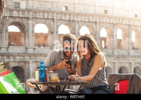 Glückliches junges paar Touristen mit Smartphone sitzen an der Bar Restaurant vor dem Kolosseum in Rom bei Sonnenuntergang mit Kaffee Einkaufstaschen lächelnd Stockfoto