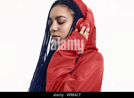 Portrait von wunderschönen afrikanischen brunette teenage Mädchen mit Dreadlocks tragen einen roten windbreaker Musik hören über kabellose Kopfhörer auf dem Bolzen isoliert Stockfoto