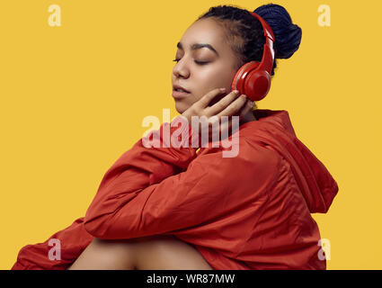 Portrait von wunderschönen afrikanischen brunette teenage Mädchen mit Dreadlocks tragen einen roten windbreaker Musik hören über kabellose Kopfhörer isoliert auf Vivi Stockfoto