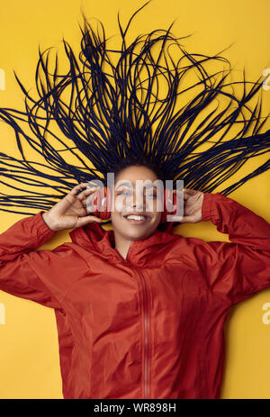 Portrait von wunderschönen afrikanischen brunette teenage Mädchen mit Dreadlocks tragen einen roten windbreaker Musik hören über kabellose Kopfhörer isoliert auf Vivi Stockfoto