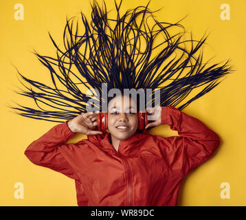 Portrait von wunderschönen afrikanischen brunette teenage Mädchen mit Dreadlocks tragen einen roten windbreaker Musik hören über kabellose Kopfhörer isoliert auf Vivi Stockfoto