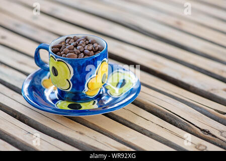 Kaffeebohnen in eine handbemalte Tasse und Untertasse auf einer hölzernen Hintergrund angeordnet. Stockfoto